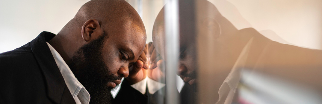 Sad looking man holding his head against a window, looking down.